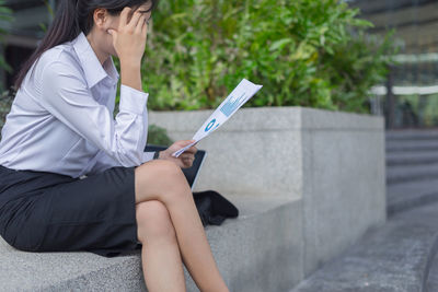 Businesswoman holding documents while sitting on retaining wall in city