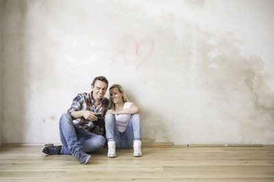 Young couple using mobile phone while sitting on hardwood floor by beige wall