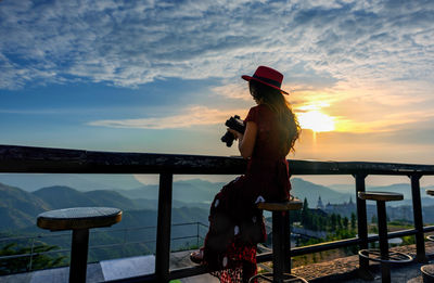 Woman standing by railing on sea against sky during sunset