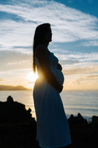 Silhouette woman standing at beach against sky during sunset