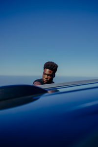 Young man standing by car at beach against clear blue sky