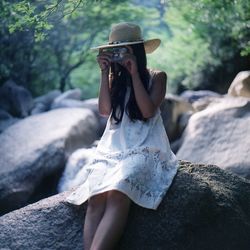 Woman photographing with camera while sitting on rock at forest