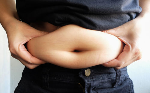 Midsection of overweight woman holding stomach fats while standing against wall