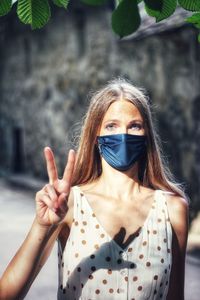 Portrait of young woman standing outdoors with coronavirus mask showing victory sign