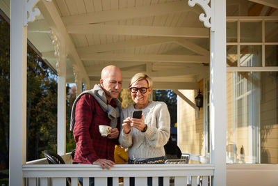 Smiling mature woman showing mobile phone to bald friend while standing on porch