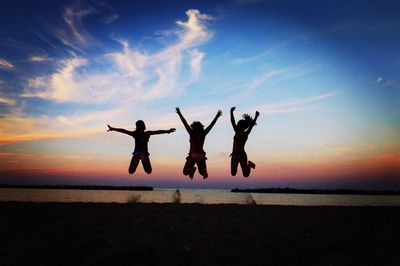 Silhouette female friends jumping at beach against sky during sunset