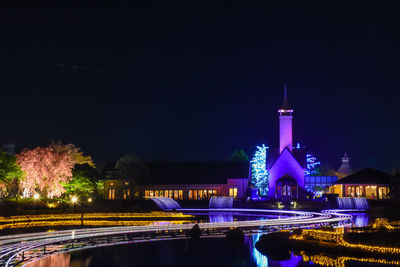 Illuminated buildings by road against sky at night