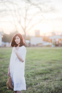 Portrait of beautiful woman holding sun hat while standing on field against sky during sunset