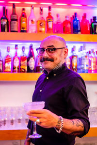 Confident stylish barkeeper with glass of cold alcoholic cocktail standing against shelves with various drinks