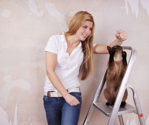 Smiling beautiful woman playing with siamese cat on ladder against wall