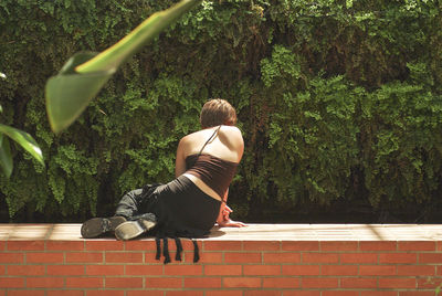 Rear view of woman sitting on retaining wall against tree