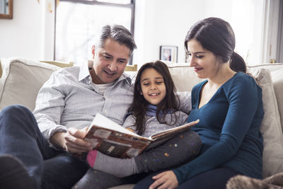 Girl with father and mother reading book while sitting on sofa at home