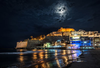 Illuminated city by sea against sky at night