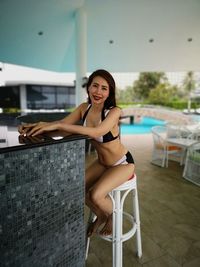Portrait of woman wearing bikini while sitting on stool at poolside