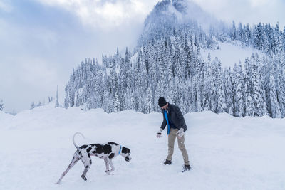 Full length of mature man playing with dog on snow field against sky