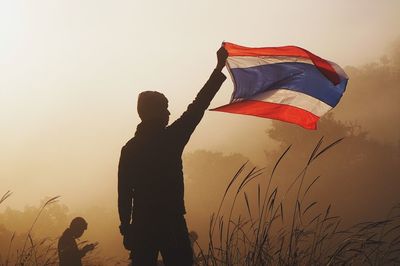 Rear view of man holding thai flag against sky during sunset