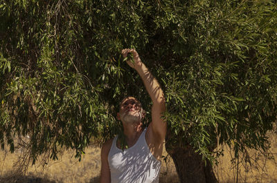 Adult man in white tank top and jeans standing with olive tree in summer