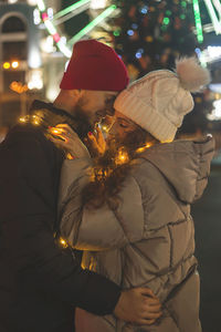 Loving boyfriend and girlfriend are hugging happily near a big city christmas tree with bokeh lights