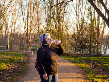 Senior woman drinking water from bottle in park during sunset