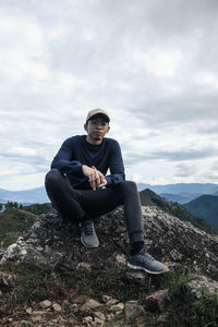 Young man sitting on rock against sky