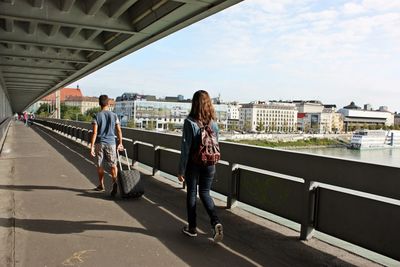 Full length rear view of people walking on footbridge in city against sky