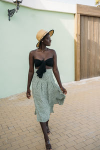 Young woman wearing hat walking against green wall at footpath