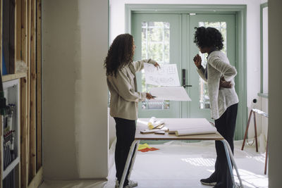 Side view of client and female architect comparing blueprints while standing near table at under construction apartment
