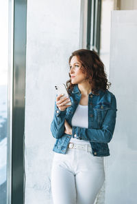 Portrait of charming young woman in jeans jacket with mobile phone near window in modern building