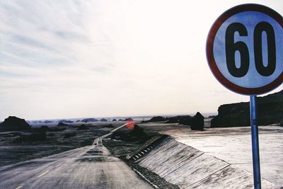 Speed limit sign by road against cloudy sky on sunny day