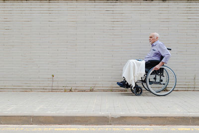 Side view of gray haired aged male in wheelchair riding along pavement in city