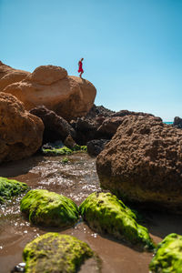 Full body side view of young barefoot female in stylish red summer dress walking on rocky coast near sea in sunny day