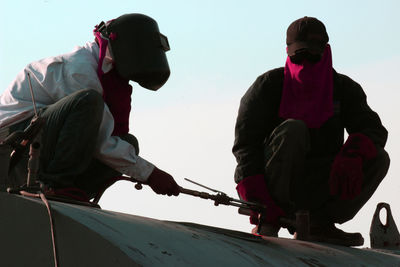 Manual workers crouching on metal while working against sky