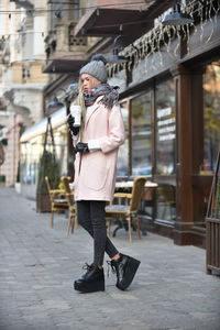 Woman in warm clothes drinking coffee while standing on sidewalk against building