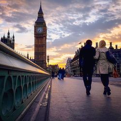 Rear view of couple walking on westminster bridge by big ben against sky during sunset