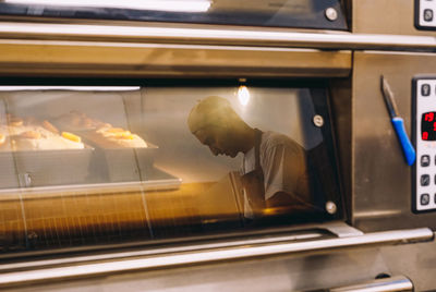 Side view of male baker in uniform standing in kitchen and reflected in glass of oven with tasty bread