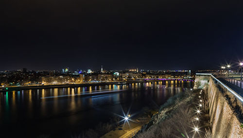 Illuminated bridge over river by buildings against sky at night