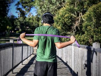 Rear view of man doing resistance band exercises while standing on footbridge.