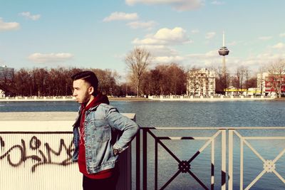 Side view of young man looking away while standing by river in city against sky