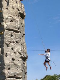 Rear view of boy hanging from rope while rock climbing against blue sky