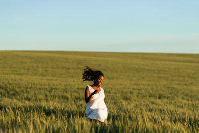 Smiling young black lady in white summer dress strolling on green wheat field while looking away in daytime under blue sky