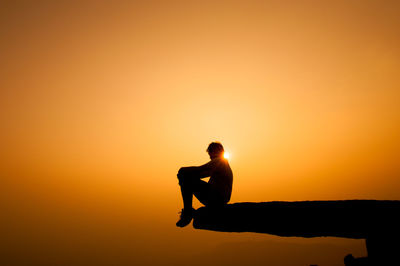 Silhouette man sitting on cliff against sky during sunset
