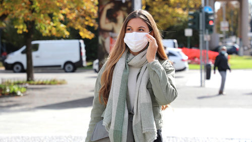 Smiling young woman wearing mask talking on mobile phone in city