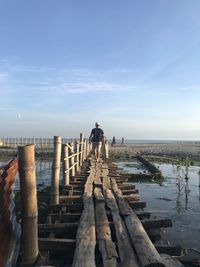 Rear view of man walking on pier over river against sky