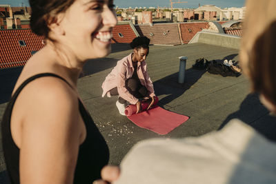 Young female folding exercise mat while friends talking after yoga on rooftop during sunrise