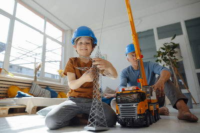 Smiling boy imitating as engineer holding electricity pylon model with grandfather in background at home