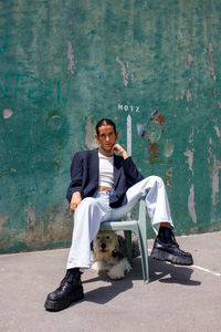 Full body of fashionable young ethnic female with dark hair in stylish outfit sitting on stool with adorable fluffy old english sheepdog under seat on weathered wall on sunny day