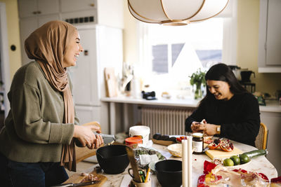 Happy young woman in hijab preparing food with friend sitting at table in kitchen