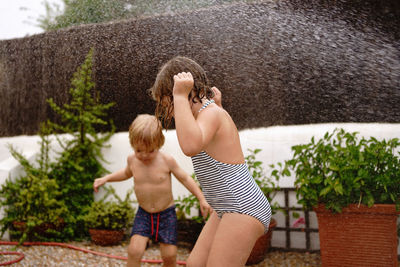 Side view of cheerful shirtless little boy pouring water from hose on sister in swimsuit while playing together in yard on summer day
