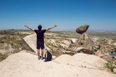 Guy in a black t-shirt stands back on a rock with a backpack spreading his hands to the sides