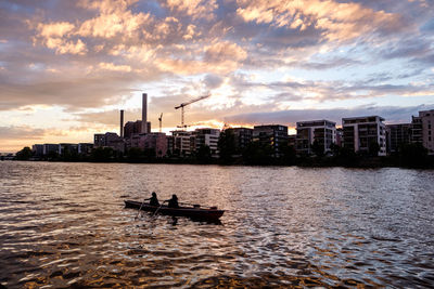 Silhouette boat in sea by buildings against sky during sunset